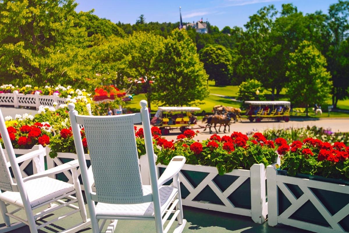 Front porch of Mackinac Island's Grand Hotel with white rocking chairs and red flowers, overlooking green grass, trees, and horse-drawn carriages.