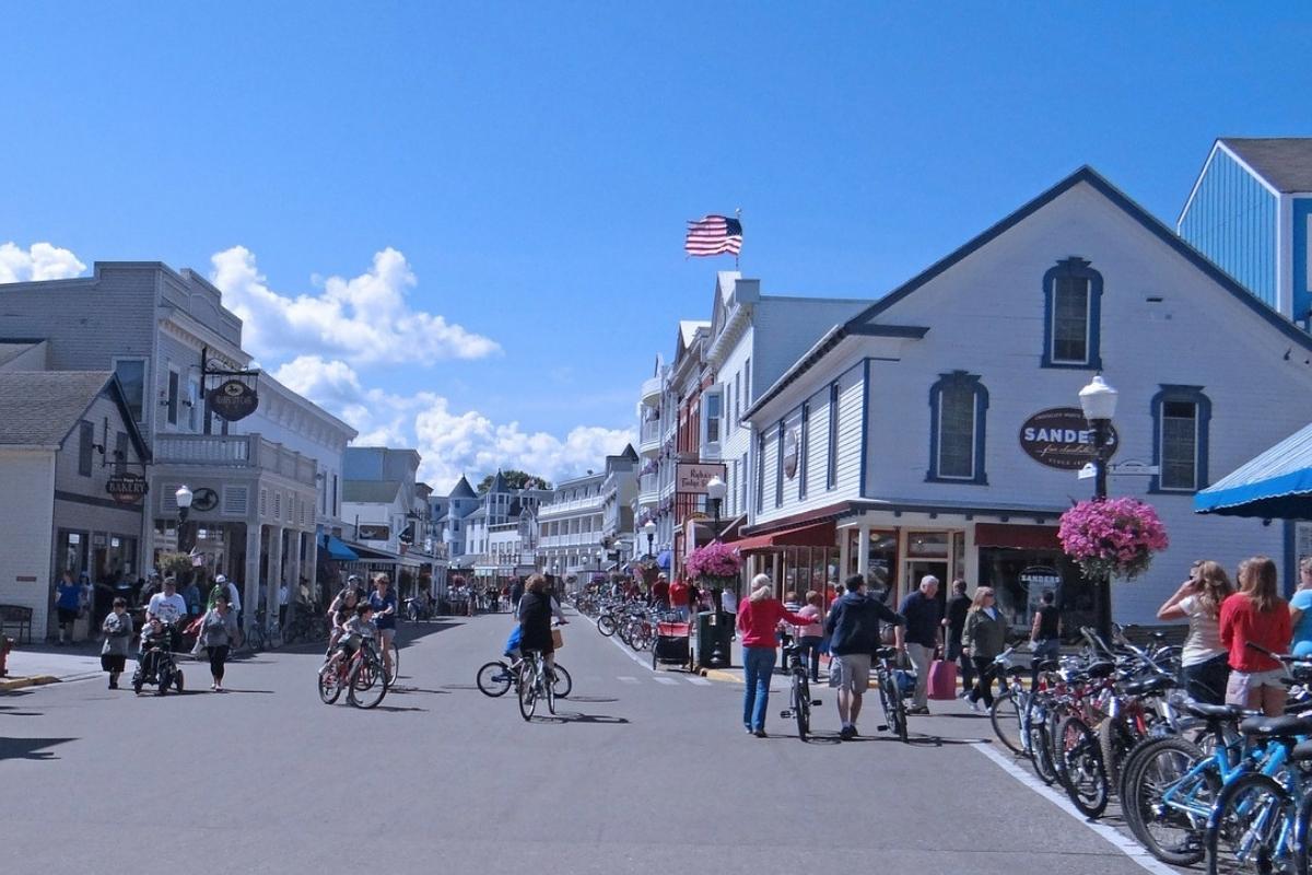 Downtown street on Mackinac Island with white wooden shop buildings, hanging pots of pink flowers, bicycles, people walking, and an American flag waving against the blue sky with white clouds