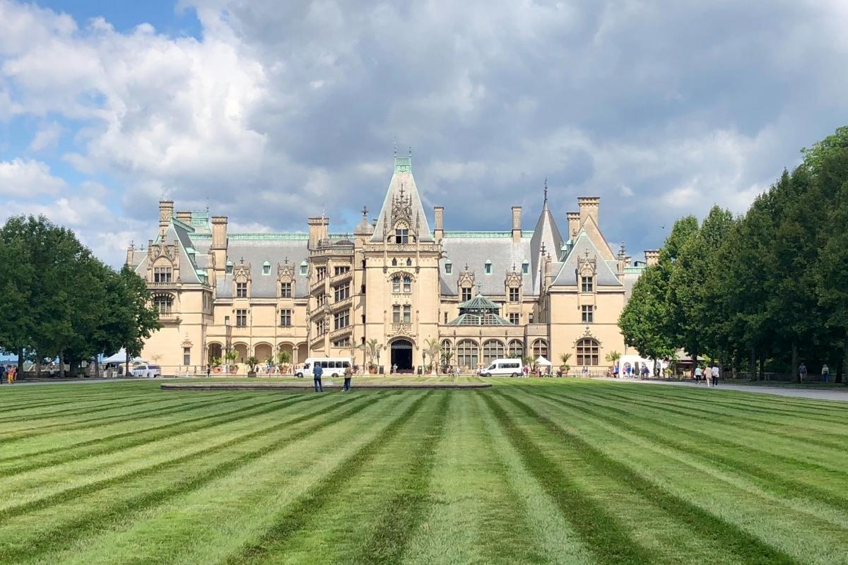 Large Châteauesque Revivalist mansion with green lawn in front, tree-lined walkways, white vans, people standing around and walking, and white and grey clouds in blue sky overhead
