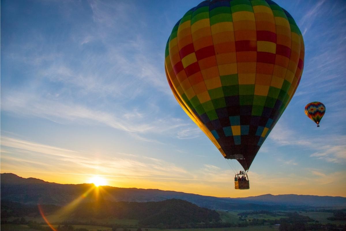 Two multi-colored hot air balloons in a blue, white, and golden-colored sky above green valleys and hills as the sun comes up.