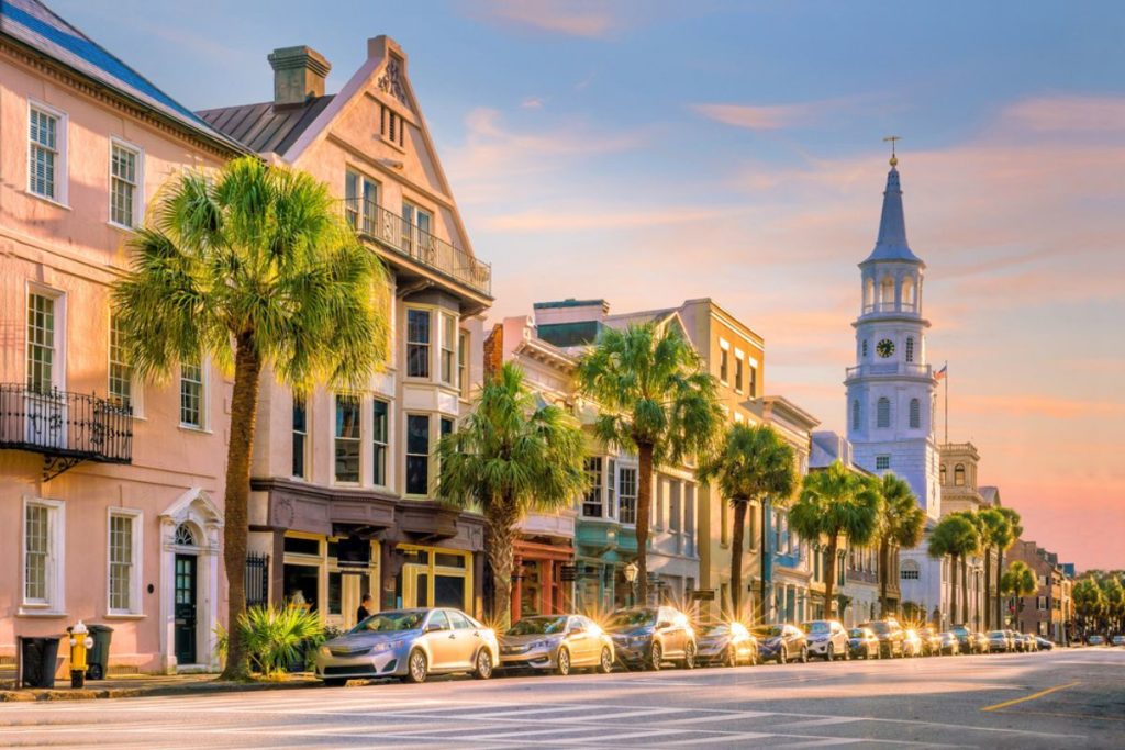 Row of historic buildings behind palm tree-lined sidewalk and parked cars, with tall church steeple in background and pink blue sunset sky overhead