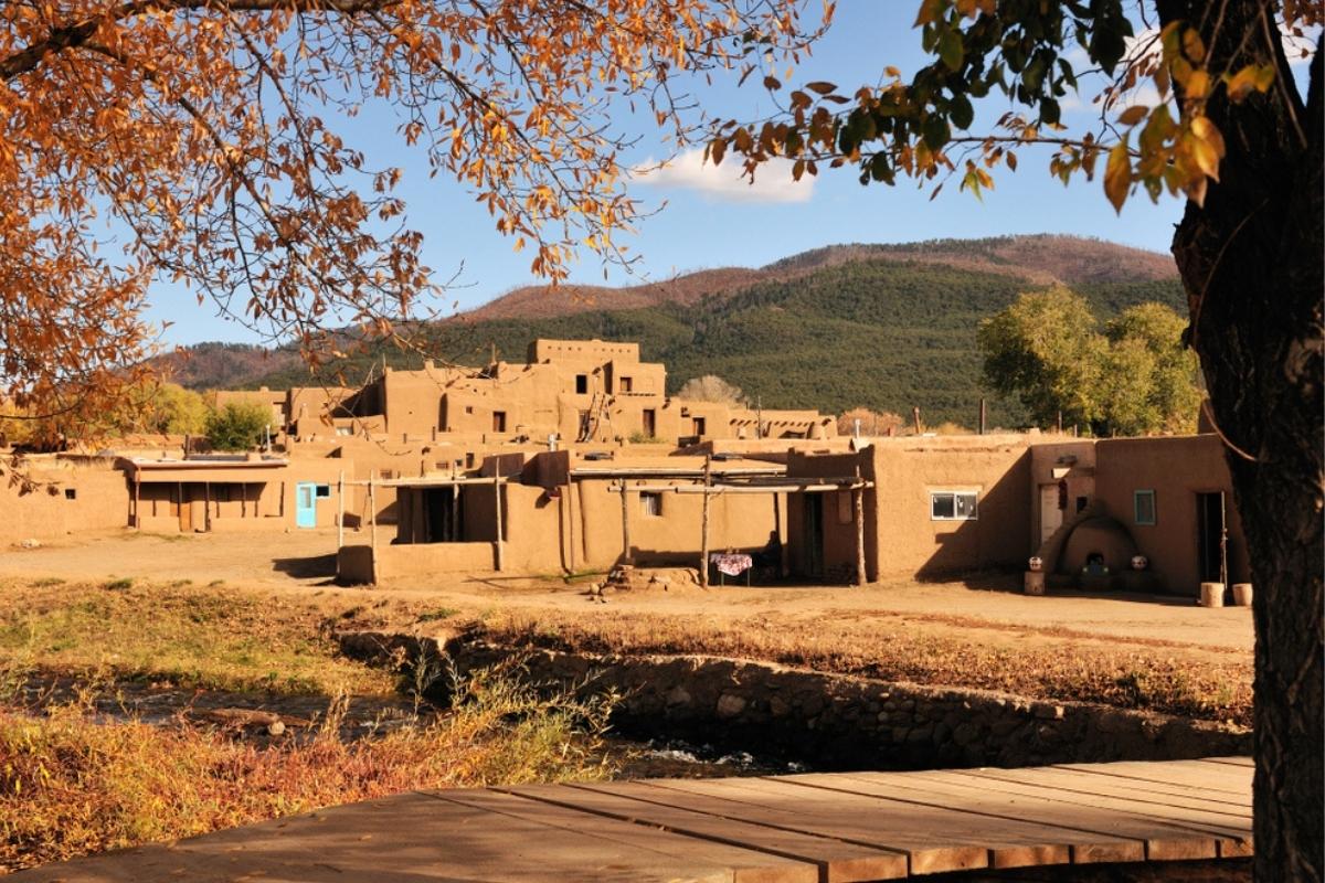 Taos pueblo with brown grass and a brown wooden bridge in front, a few trees around, and green mountain behind