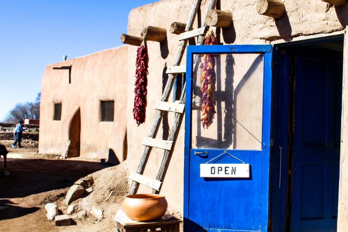 light brown pueblo building with an open blue door and a black and white "open" sign, dirt walkway alongside it, wooden ladder leaning against the wall, two bunches of red chilis hanging from wooden posts on either side of the ladder, a bronze-looking rounded pottery piece on a small wooden table beside the door and ladder, a person walking in the background, and clear blue sky overhead