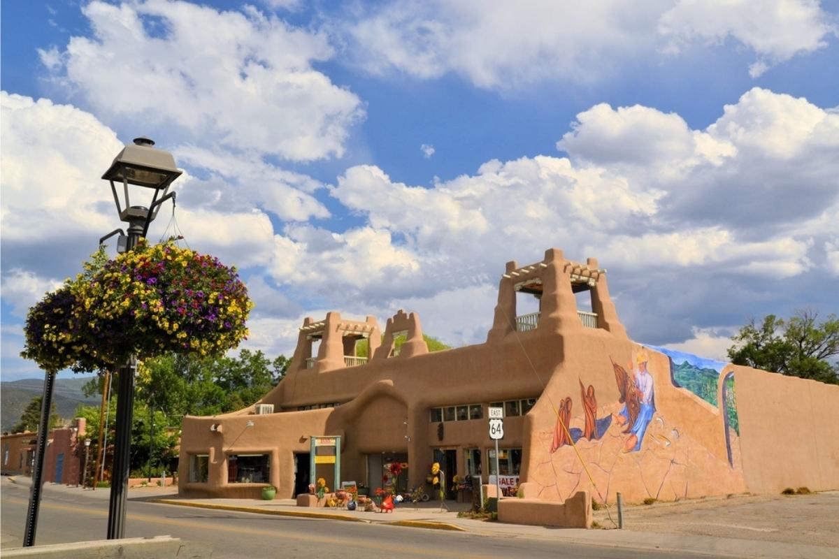 A pueblo building with mural on the side, empty street in front, surrounded by a few trees and hanging flower baskets, with blue and white cloud sky overhead