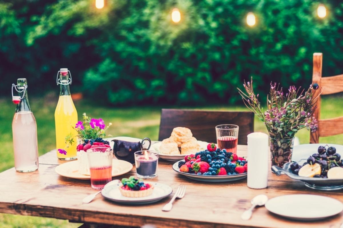 brown picnic table in backyard with berries, black cherries, bottles and glasses of juice, purple and green flowers, a white candle, and four place settings, with a string of lights overhead
