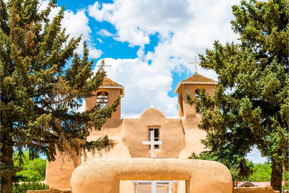 historic pueblo style mission church with small white cross, surrounded by green trees on the side and background, with blue and white cloud sky overhead