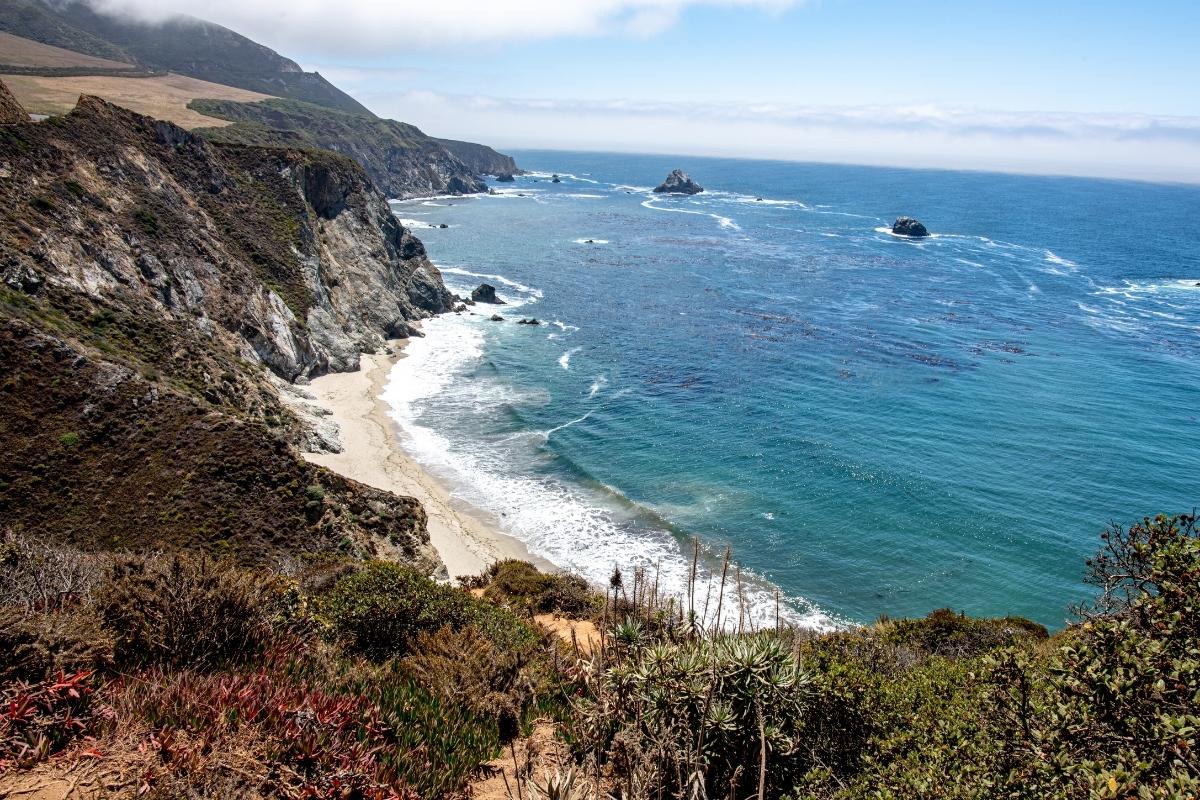 View of the water and small sandy beach along the Monterey, California coastline. Taken from Bixby Bridge, there's an expanse of blue water lined with craggy, rocky cliffs, and light blue sky with white clouds overhead.