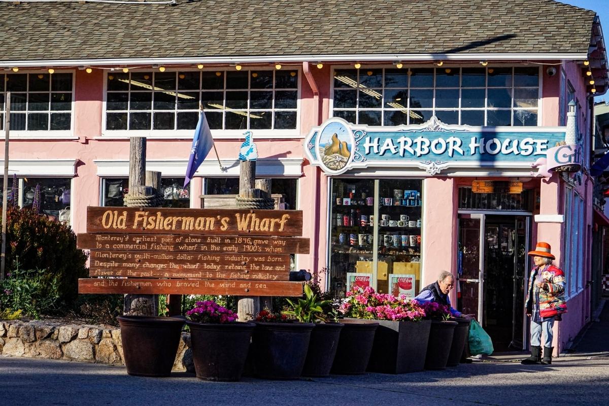 Harbor House store at Old Fisherman's Wharf in Monterey, California. The building is pink with a blue sign, store windows show shelves of mugs and other souvenirs. In front of the building, there are pink flowers, green plants, a low stone wall, and a wooden sign with historic information about Old Fisherman's Wharf. An elderly man sits outside the door of the shop holding a light blue shopping bag, and an elderly woman wearing a multicolored jacket and red hat stands nearby.