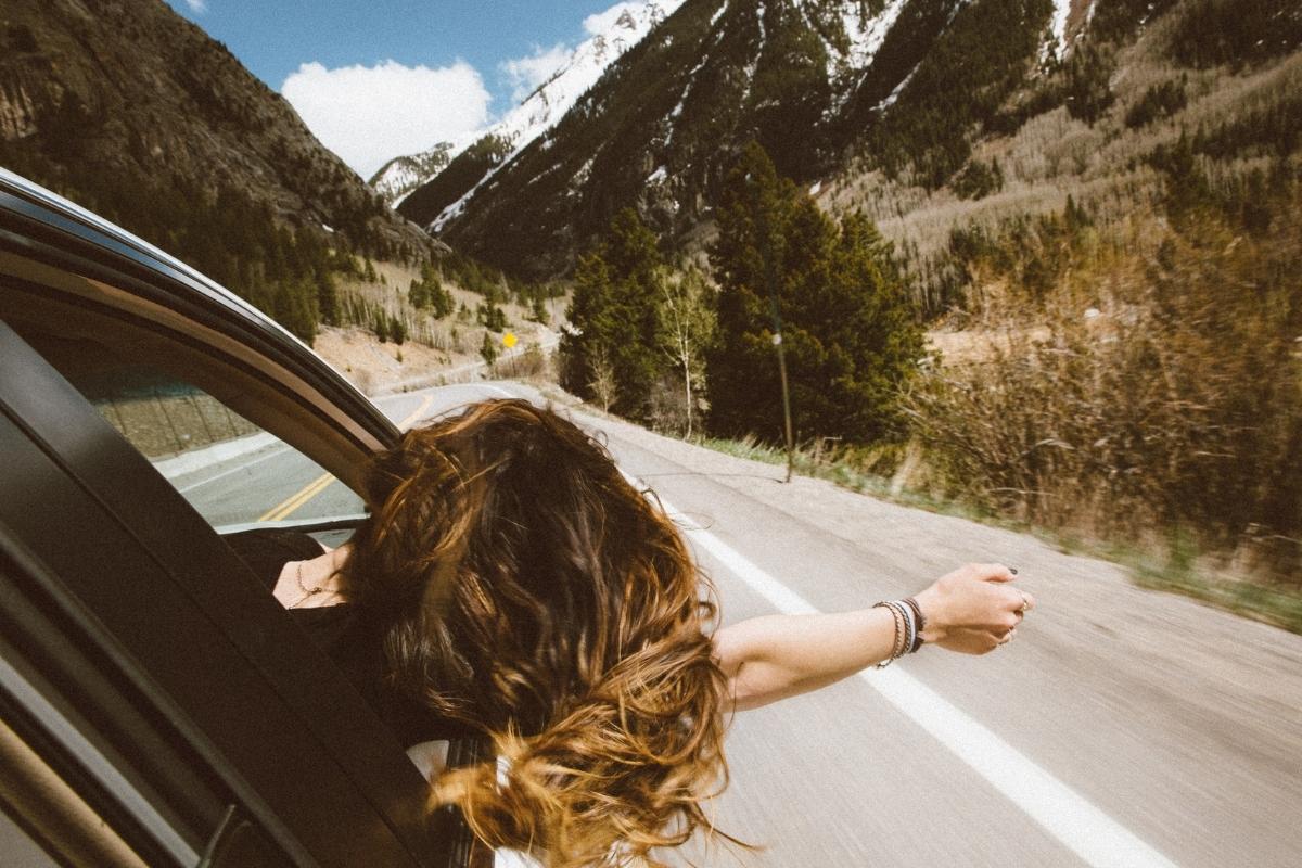 Woman with long brown hair and black shirt leaning her head and arm out the passenger side car window. Empty road stretches ahead with trees, tall grasses, and hilly landscape along the side of the road and snow-topped mountains just ahead.