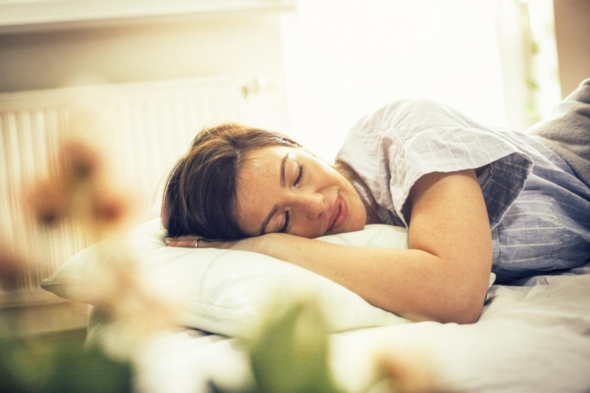 Woman with brown hair, sleeping with a smile on her face. The bedding is white, she's wearing blue pajamas with thin, white stripes, and there is a light-filled window out of focus in the background and a plant with green leaves out of focus in the foreground.