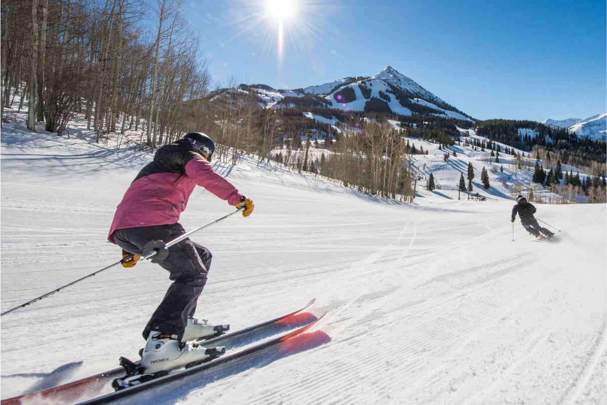 Two skiers going down a winter, tree-lined slope with sunny blue sky overhead and snow covered mountain peaks in the distance.