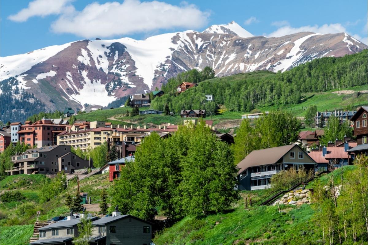 Houses and buildings on a green, tree-covered hill with snow-covered mountain in the background and a blue sky with fluffy white clouds overhead.