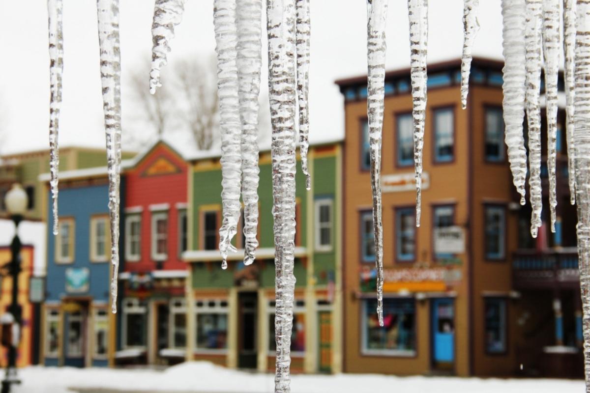 Winter street scene with a group of hanging icicles in the foreground with a row of colorful storefronts across the street in the background. The sky overhead is white and grey.
