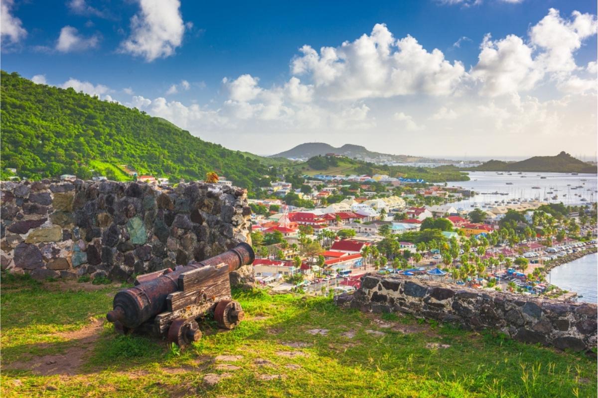 The town of Marigot on the Caribbean island of St. Martin. The view is from the ruins of Fort Louis, and shows an old cannon, the remains of stone walls, and the city and water below. The buildings of Margot have red roofs, and there are boats and green hills in the distance. Above, the sky is blue with large white clouds.