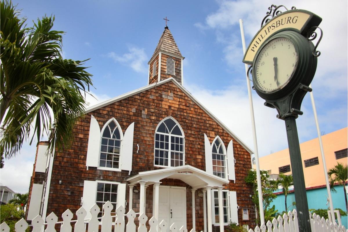 Brown and white wooden Methodist church in Philipsburg St. Maarten. There are white shutters, a white door with a small covered entrance, and a white picket fence in front. The green of a palm tree can be seen on the left side and a tan and blue buildings with other palm trees are on the right side of the church. There's a clock on a dark metal sign post in front of the church. The time is 6:30 and the word on the sign is Philipsburg. Above, the sky is blue with wispy white clouds.