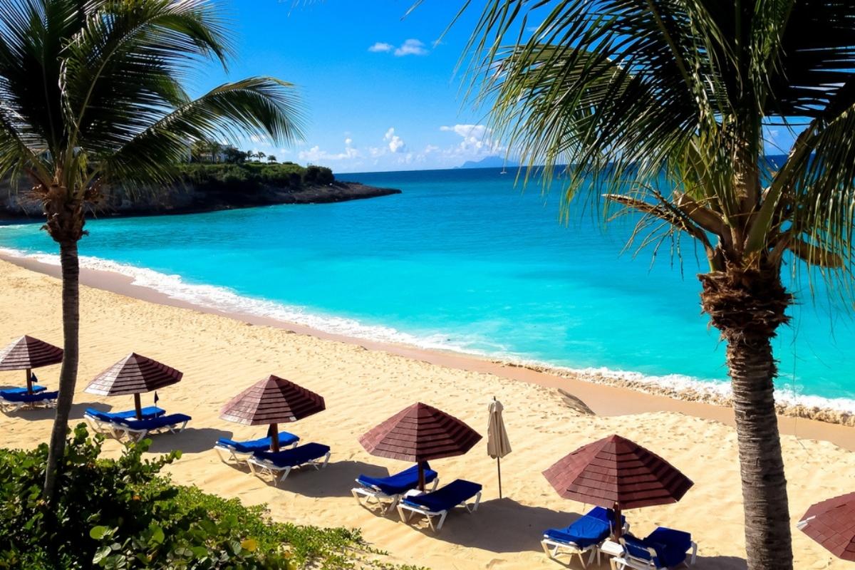 Caribbean beach with golden sand, paln trees, and five sets of blue and white beach chairs with brown umbrellas. The water is calm and turquoise with gentle white surf on the shore. There are green hills off to the side in the background and blue sky with white clouds overhead.