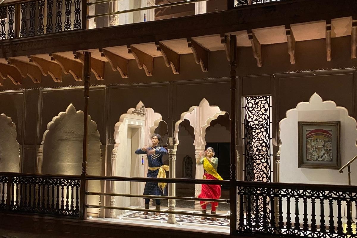 A man and a woman in traditional Indian clothes, dancing on an ornate covered balcony (with curved archways, iron railing, and stone tiles). The lighting is mostly dark except where the dancers are.