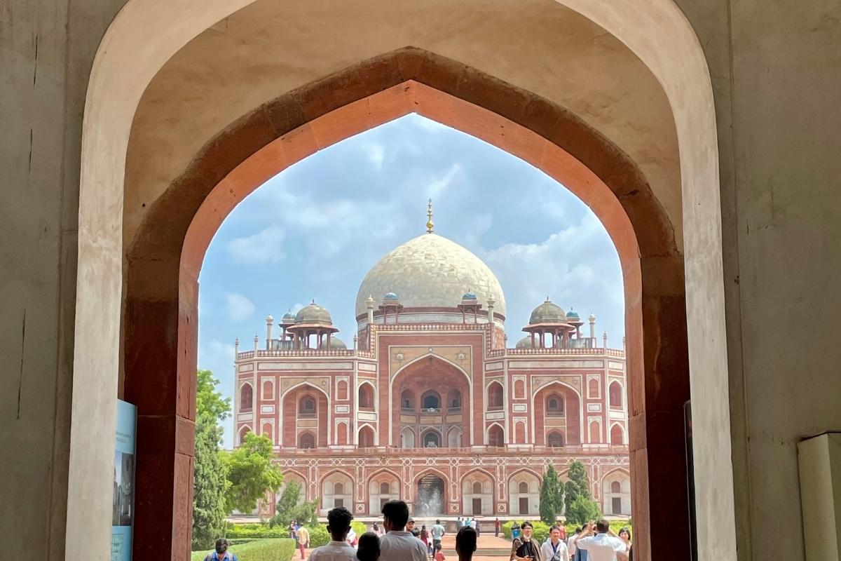 A series of curved archways opening to Humayun's Tomb set within green landscaping, with people in front and blue and white sky overhead.