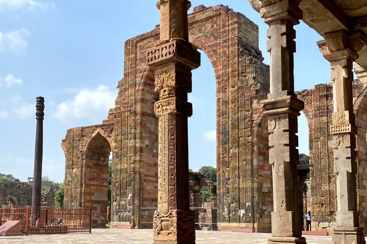 Stone columns and arches at the Qutub Minar in India. Stone is varied shades of brown, the sky is blue with soft white clouds, and green tree tops can be seen in the background.