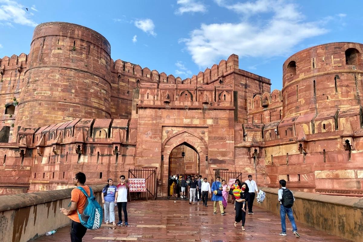 The red stone Agra Fort, with walkway, arched entrance, and rounded turrets. Visitors are on the walkway coming out of the fort.