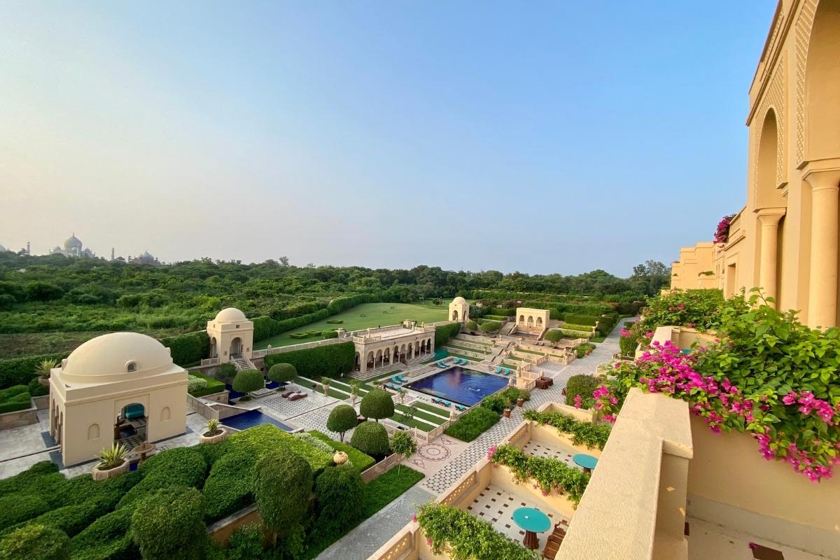 Exterior of the hotel and courtyard, with stone patios and domed outdoor buildings, tile walkways, blue pool with lounge chairs, and lush green gardens and landscaping surrounding it. In the distance, you can see the top of the Taj Mahal.