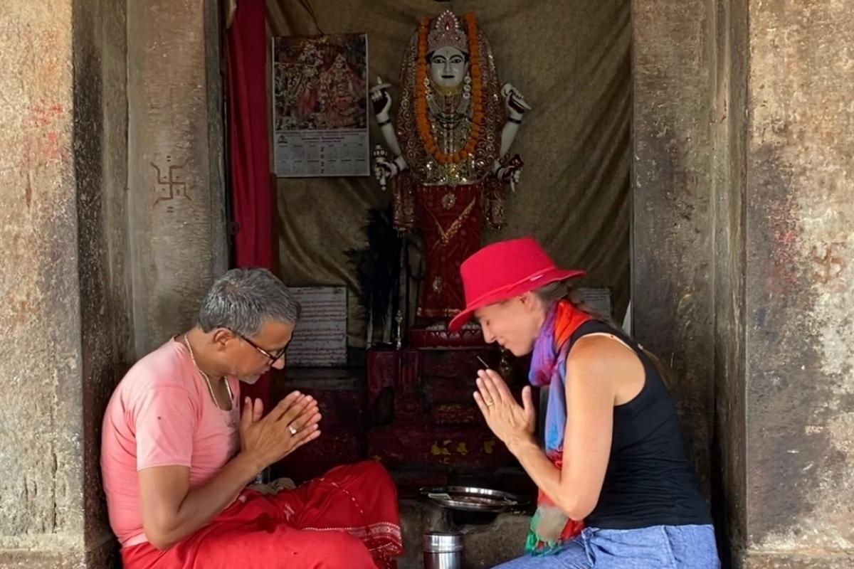 Melissa receiving a happiness blessing from the holy man at the Harshat Mata temple. They are seated across from each other, with eyes closed and hands in prayer position, on a stone step in front of the temple. A colorful statue of the goddess is behind them and the ancient Hindu symbol for well-being is on the wall.
