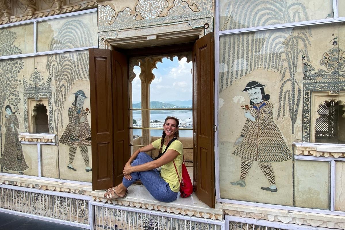 An interior muraled wall at the City Palace of Udaipur, with a small window seat opening to views of the city and the Lake Palace from a scalloped window. Melissa (the author of the post) sits on the window seat, smiling and wearing a yellow shirt, blue pants, gold-colored sandals and a red bag.