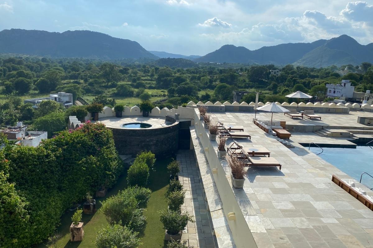 Wide shot of the swimming pool at Devigarh in Udaipur, India. Stone patio with a few loungers, umbrellas, and plants surround the pool to the  right of the photo. And expansive views of the surrounding green landscape, with lush mountains in the distance and blue and white skies overhead fill the rest of the photo.
