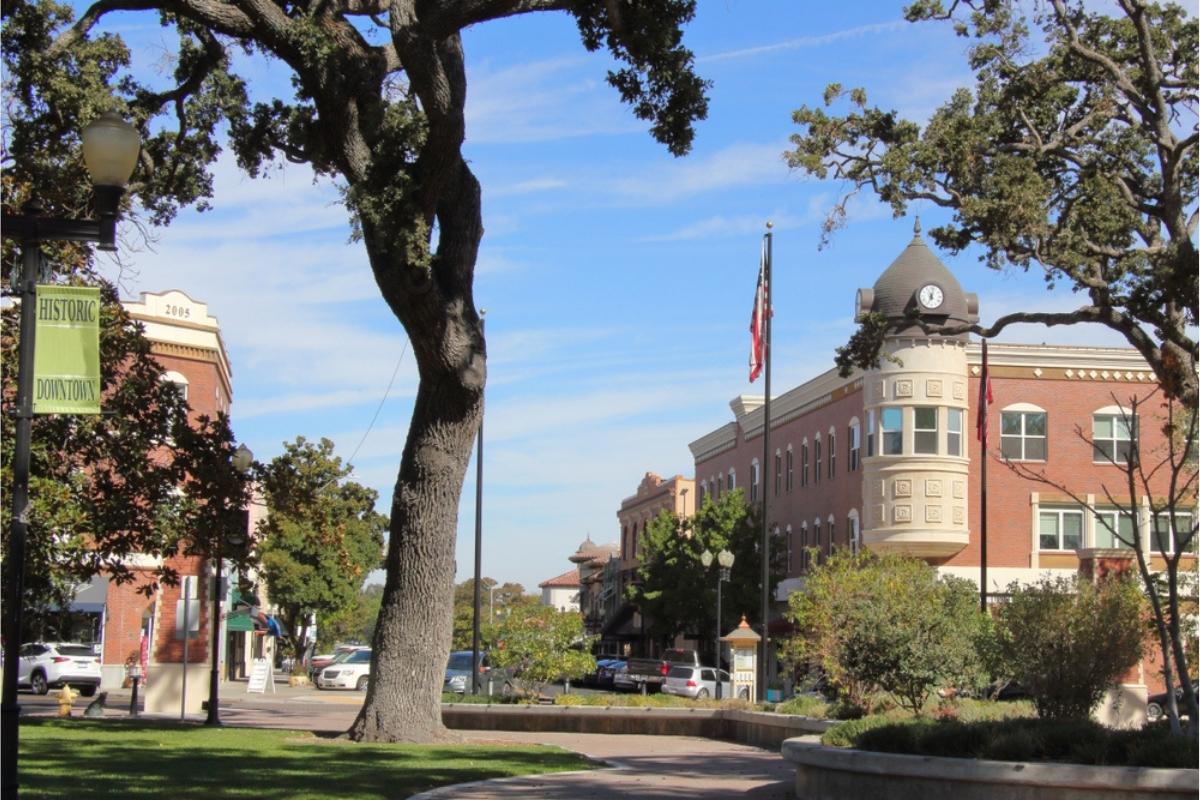 Downtown Paso Robles view from shaded park with a few trees in the foreground; buildings, cars and flag in the background; and blue sky overhead.