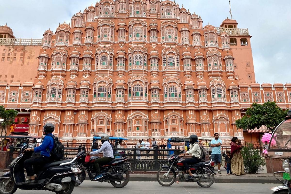 Street view of the Hawa Mahal in Jaipur. The building is large with dark pink walls and rows of large arched windows. In front of the building, people are walking, riding scooters and motorbikes, or in rickshaws on the paved street.