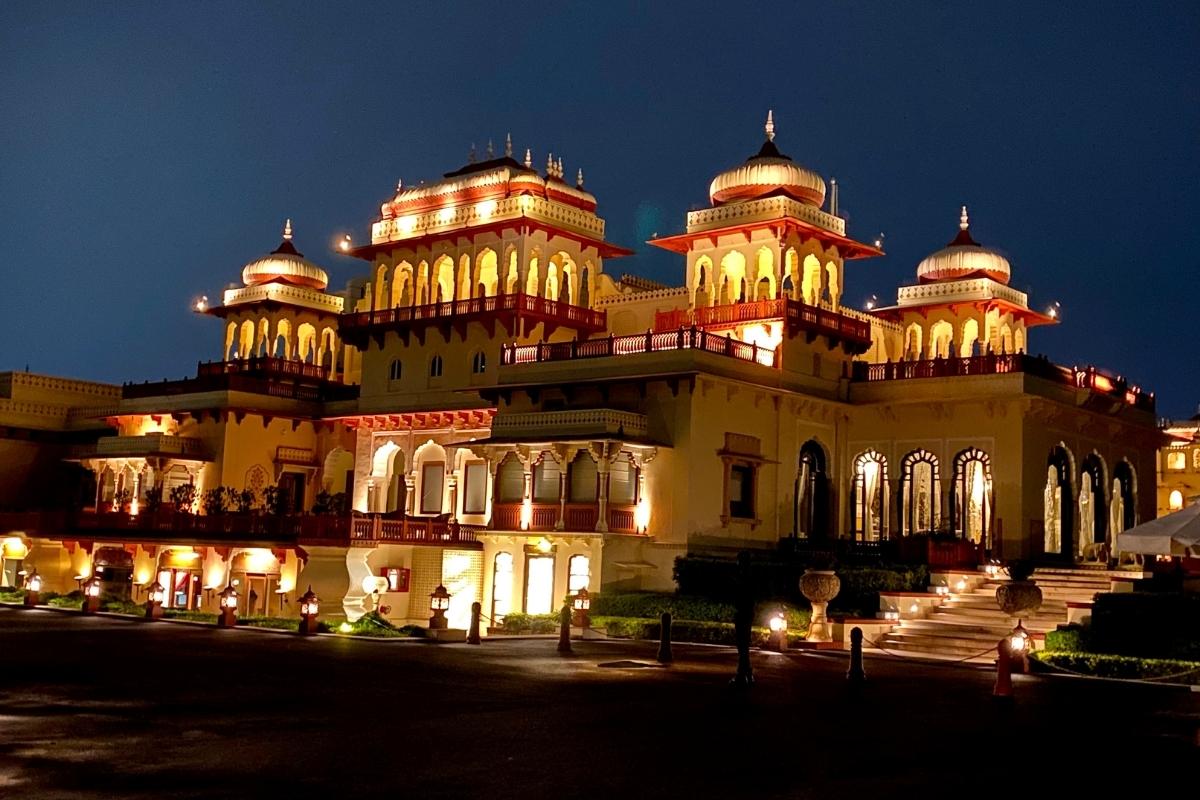 Queen's Palace in Jaipur at night. The palace is lit with bright interior lights and rows of smaller lights outside lining the walkway and stairs. The palace is cream colored with red details, three domed sections on the roof, scalloped archways and multiple balconies, set against a dark blue night sky.