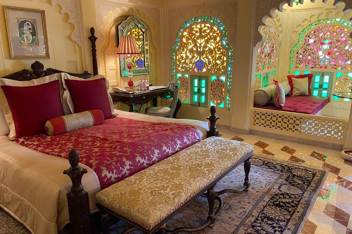 Ornate bedroom with Indian-style architecture, queen bed, cream-colored walls, stained glass windows, window seat, side table with lamp and chair, and rich, jewel-toned linens and pillows.