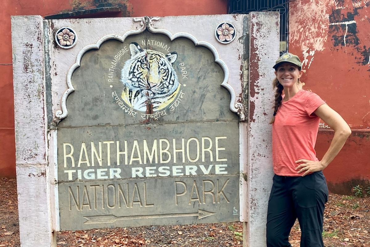 Tall wooden entrance sign at the Ranthambore National Park with a painting of a tiger at the top, scallop details, and distressed paint. The blog post author, Melissa, is standing beside the sign, smiling and wearing a light red tshirt, dark pants, and a green cap.