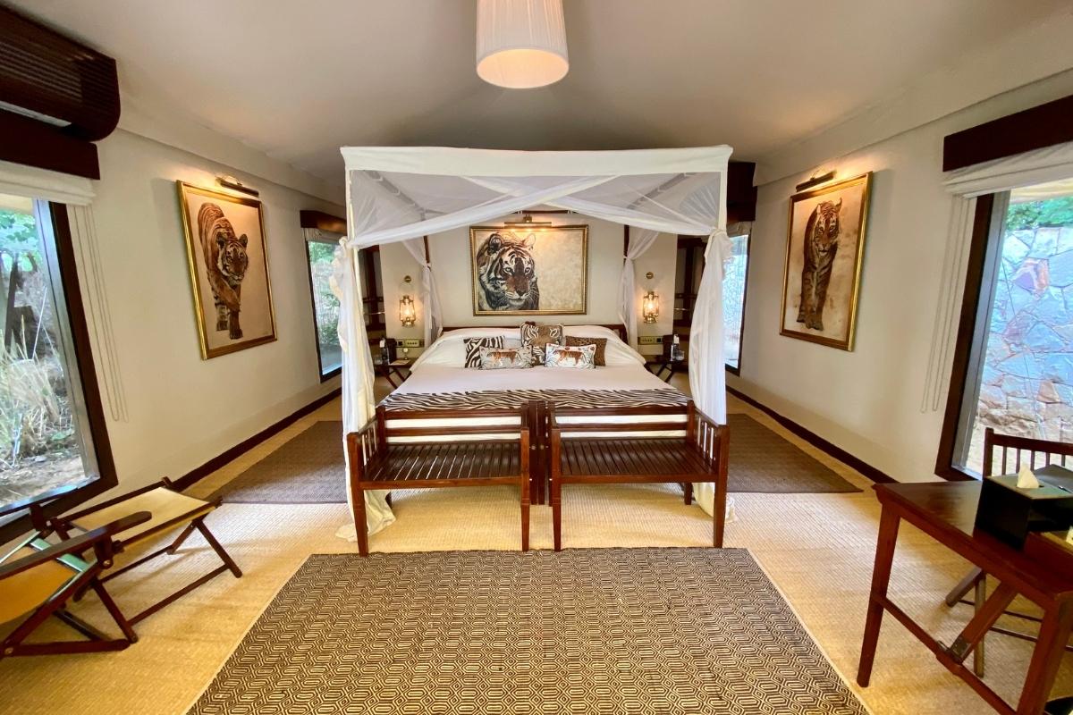 Interior of a large luxury safari tent. There is a queen size four-poster bed with mosquito netting, rugs on teh floor, paintings of tigers on the walls, and wooden chairs and a wooden desk. The lighting is soft and there are four large windows showing the landscape outside.