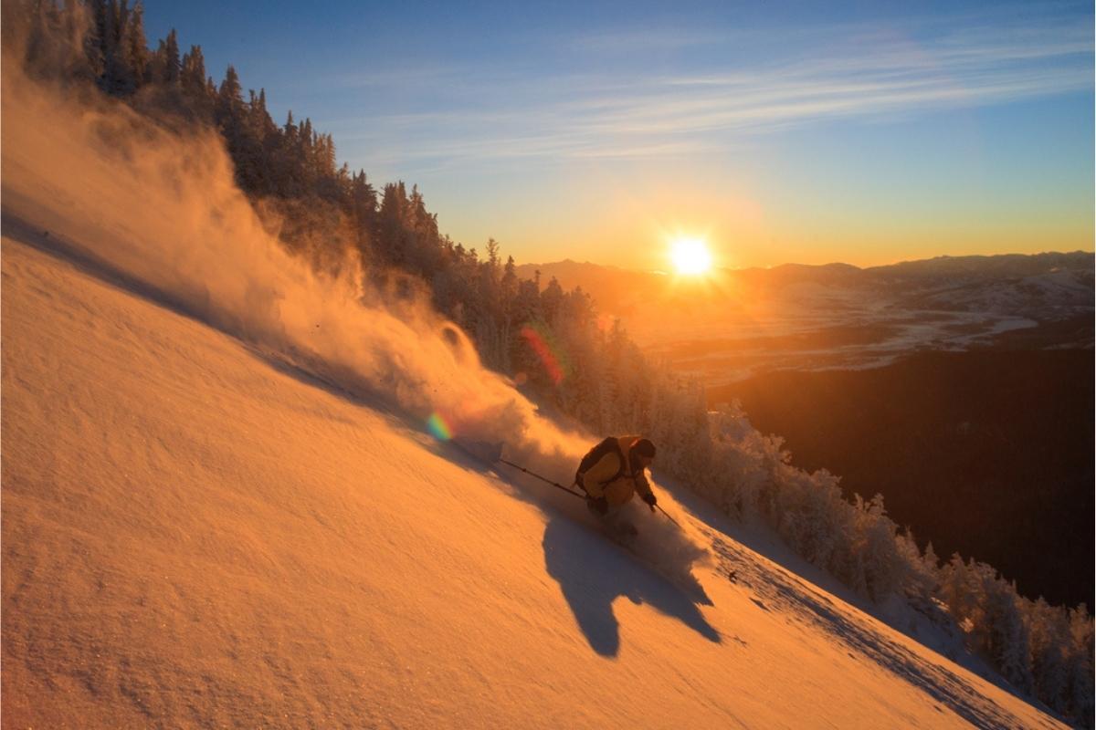 Skier going down steep, snow-covered mountain at sunrise. Powdery snow flies behind him, snow-covered trees line the mountain behind him, and the sun rising above the mountains in the far distance casts an orange tint to everything below.
