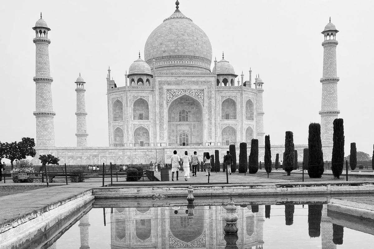 Black and white photo of the Taj Mahal, with stone walkways lined with tall narrow hedges, pool of water in front, and people walking towards the building with their backs to the camera.