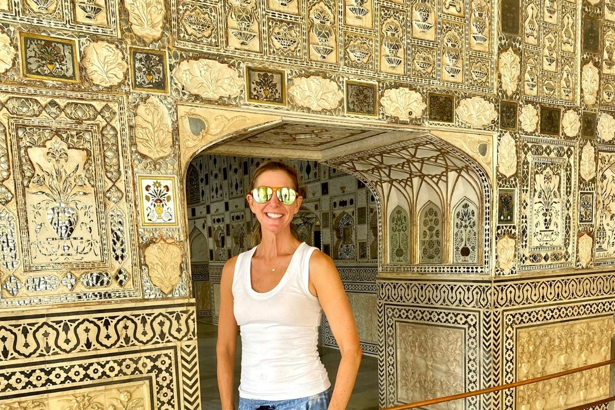 The blog post author, Melissa, smiling and wearing a white top and sunglasses, standing in front of one of the gold and silver walls at the Amber Fort.