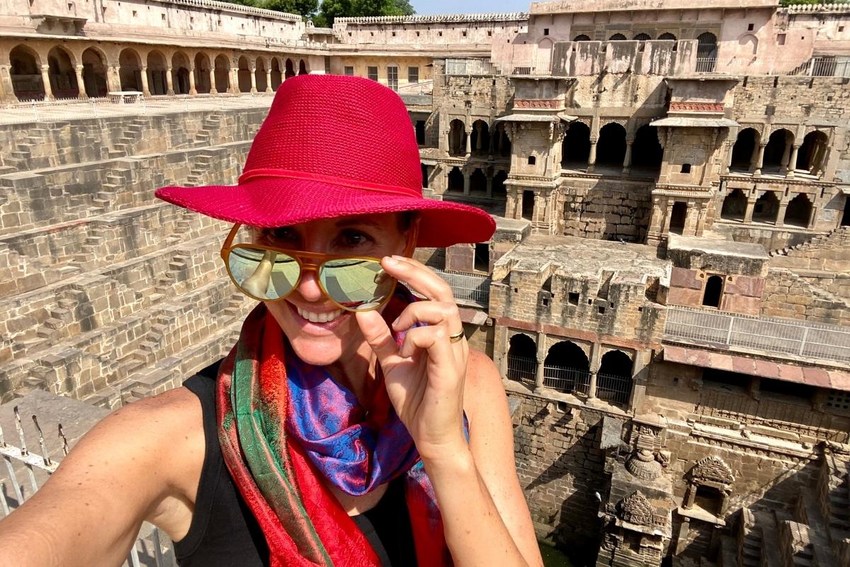 Selfie of Melissa in front of ancient stepwells on the way to Agra, India. She's smiling and wearing a red/magenta hat, sunglasses, a black top and colorful scarf.