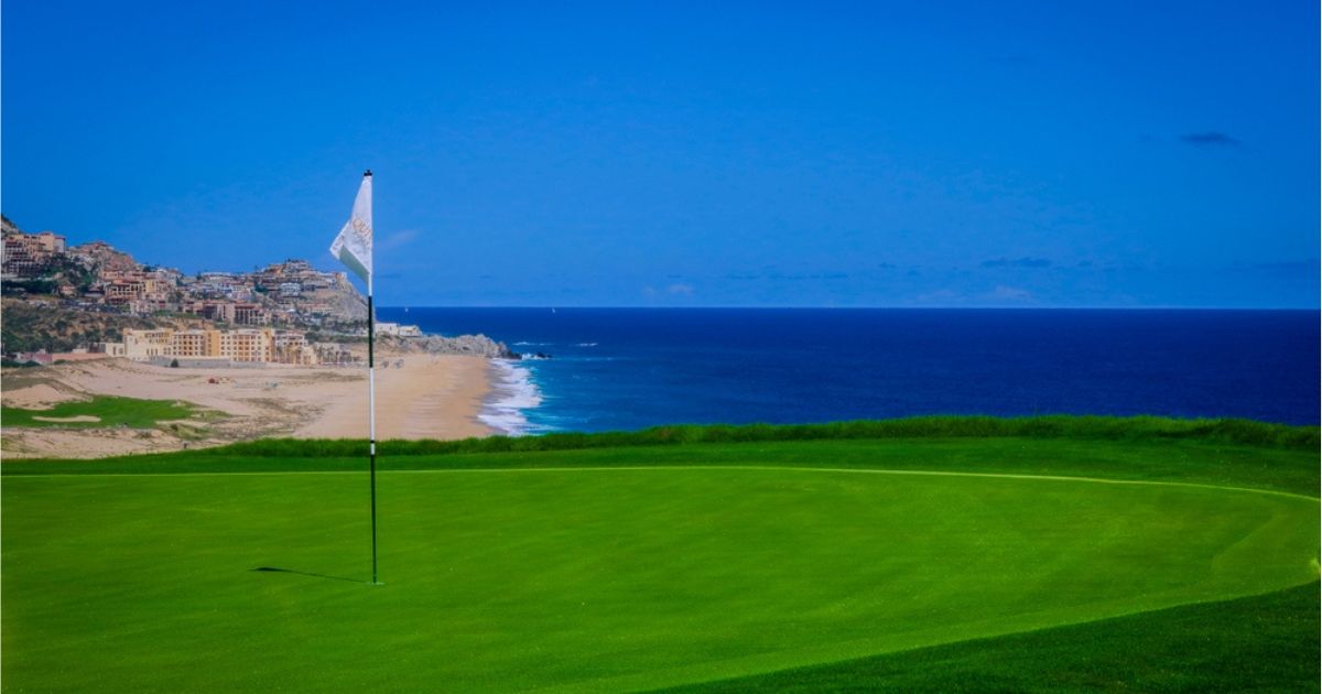 Flag next to hole on the greens at a Cabo San Lucas golf course overlooking dark blue water, light blue sky, and golden sand beach and resort buildings in the background.