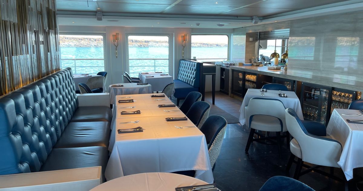 Onboard dining room with blue, white, and brown decor, tables and booths, and large windows with views of the water outside