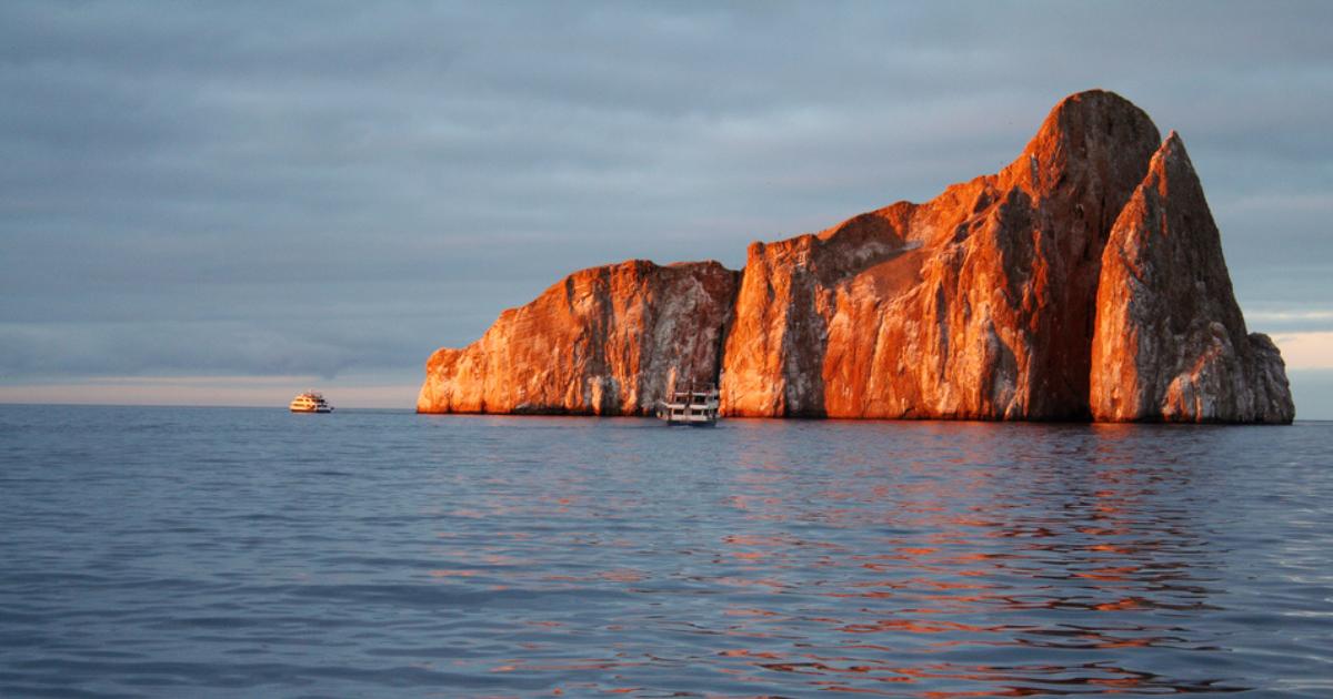 Kicker Rock/Leon Dormido at sunset, with shades of orange and red, blue sky and water, and two boats nearby