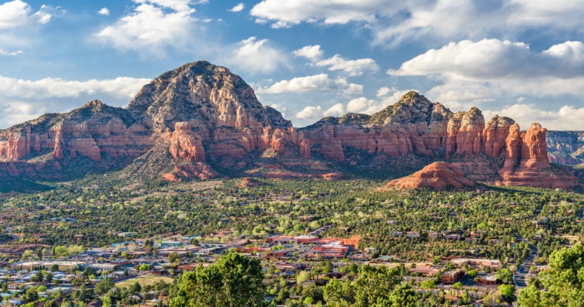 Aerial view of Sedona, set within green and brown landscape with red rock mountains in the background and blue sky and white clouds overhead