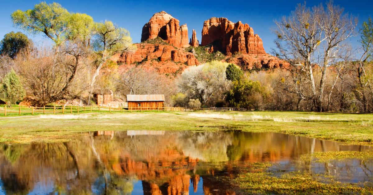 View of Sedona's Cathedral Rock in the background, with trees, green grass, a small wooden building, and pond in the foreground and clear blue sky overhead