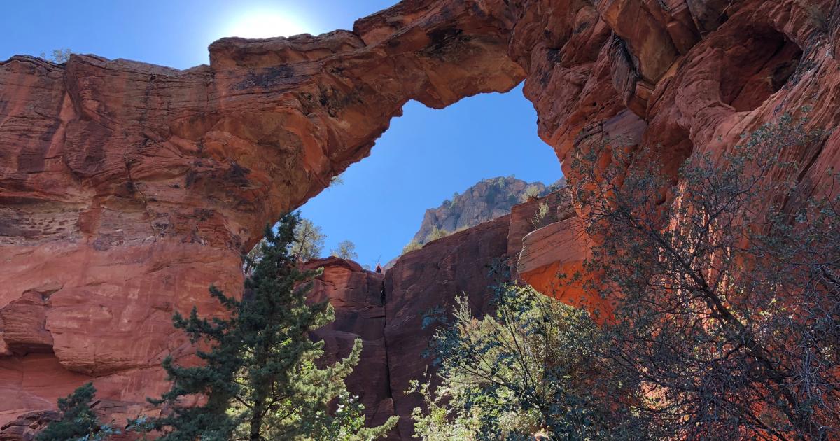 View from beneath the towering red rocks of Devil's Bridge in Sedona, with green trees in the foreground and blue sky overhead