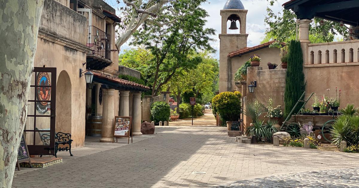 Walkway, storefronts, potted green plants, and bell tower at Tlaquepaque Village in Sedona, AZ, with green trees and light blue sky in the background