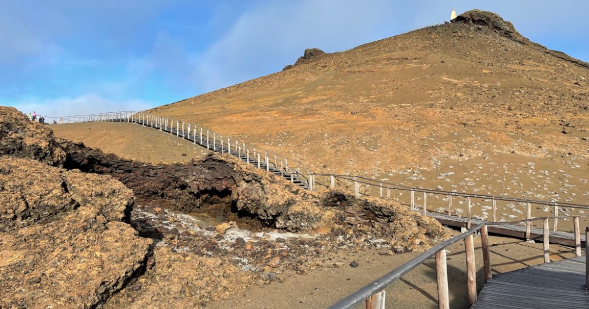 Long wooden boardwalk with railing going up brown, rocky hills to the top of Bartolomé Island, with a blue sky overhead.