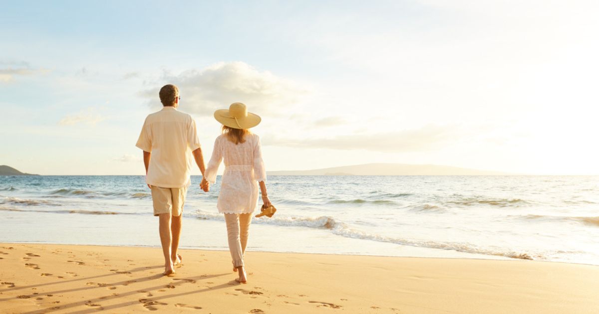 Couple wearing white, walking on the beach at sunset with calm water, golden sand, and light blue sky