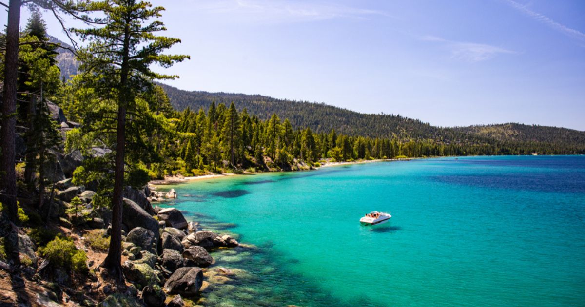Aerial view of a boat on the blue green water in Lake Tahoe, surrounded by rocky coastline, evergreen forest, and rolling hills in the background