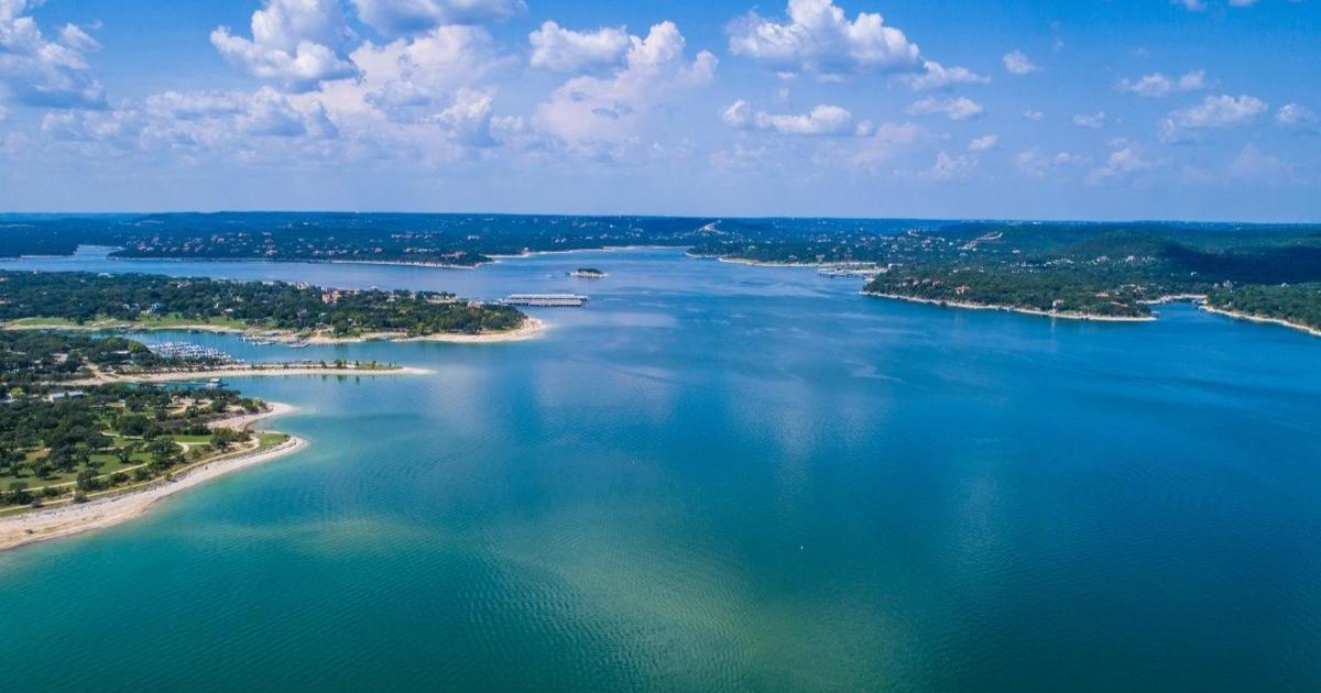 Aerial view of the deep blue green water of Lake Travis, lined with beaches, trees, and boat docks in the distance and blue sky and white clouds overhead