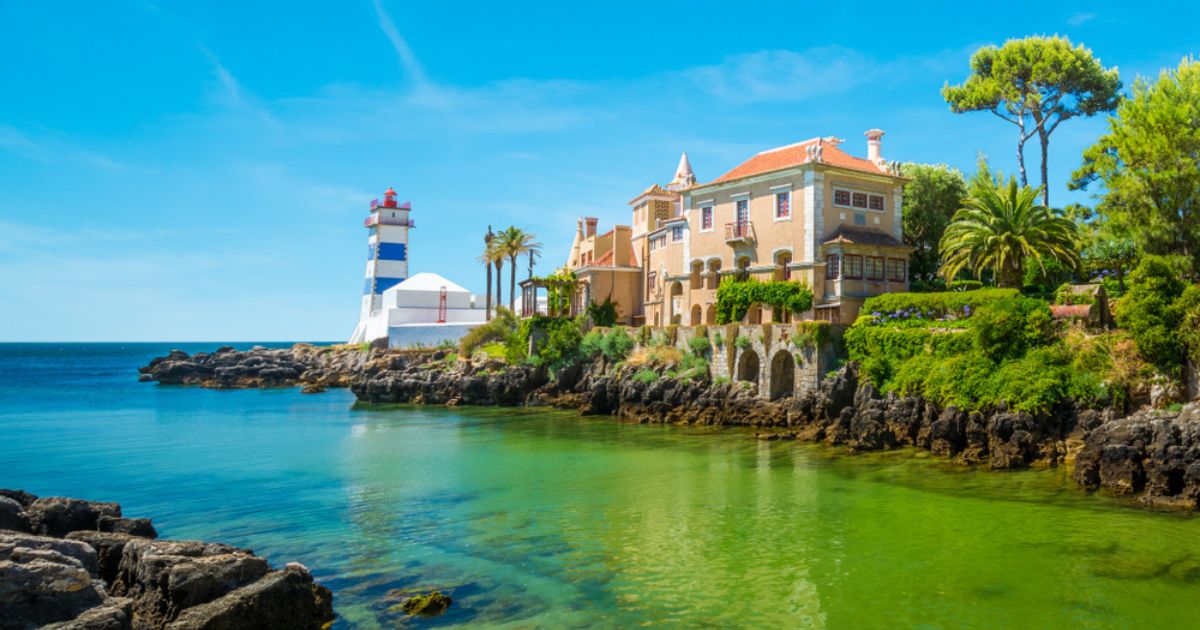The Santa Marta Lighthouse in Cascais, near Lisbon. The light brown and white building next the blue and white striped lighthouse is surrounded by lush, green trees and landscaping. Blue-green water is in the foreground and background and the sky overhead is a deep blue.